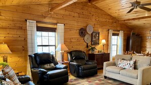 A cozy cabin interior with wooden walls, two leather chairs, a white sofa, and a patterned rug.