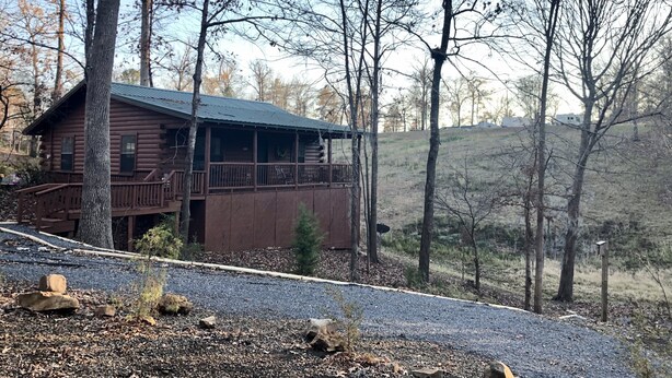 A rustic log cabin with a balcony, surrounded by trees and a gravel driveway.