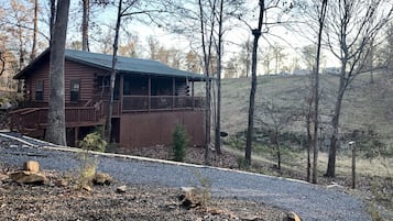 Un chalet rustique en rondins de bois, doté d’un balcon, entouré d’arbres et bordé d’une allée en gravier.