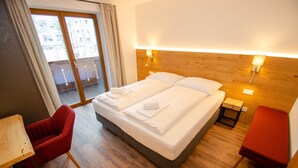 Hotellihuone, jossa on suuri sänky, puinen pääty, kaksi yöpöytää, punainen tuoli ja näkymä ulos liukuoven kautta.