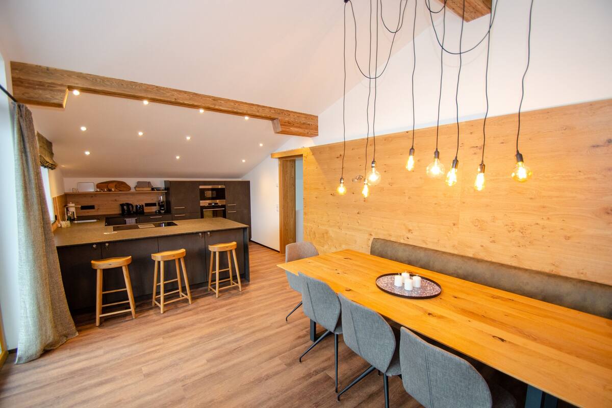 Une cuisine moderne agrémentée d’éléments en bois, un coin repas avec une table en bois et des tabourets de bar, et un luminaire suspendu.