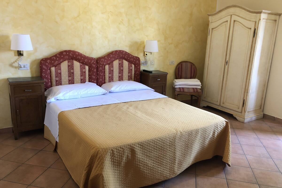 Una cama bien hecha con cubrecolcha color beige, dos mesitas de noche con lámparas, una silla de madera y un armario.