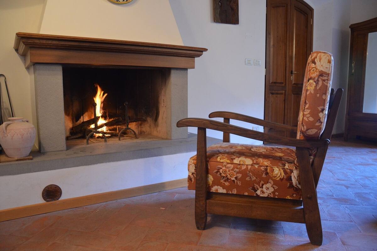 Una sala de estar con chimenea, una silla de madera tapizada con motivos florales y un aparador de madera.