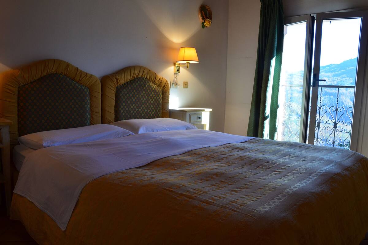 Habitación de hotel con una cama grande, una lámpara de noche, una mesita auxiliar y vistas al exterior a través de una ventana.