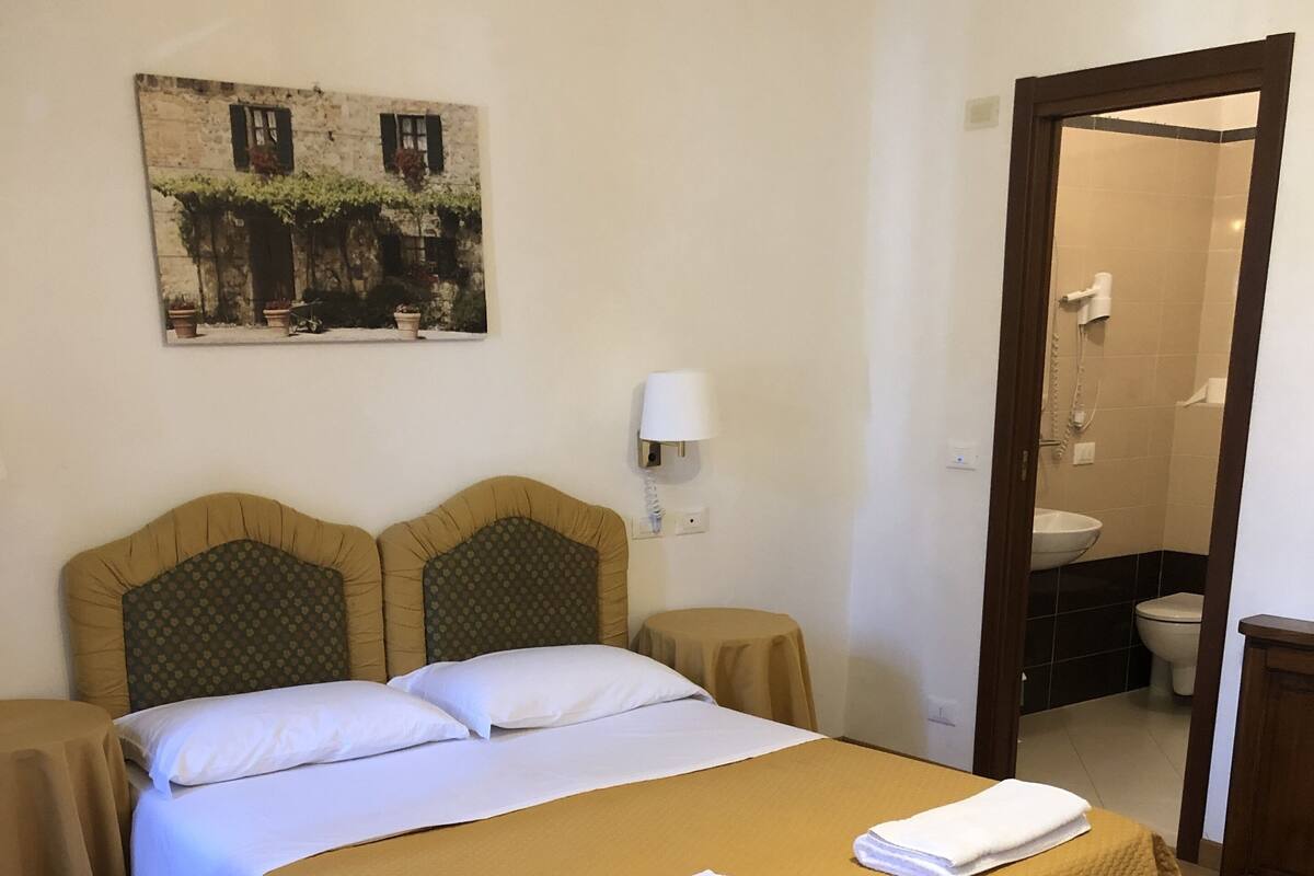 Habitación de hotel con cama, mesita de noche, espejo, baño visible a través de una puerta abierta y un cuadro colgado en la pared.