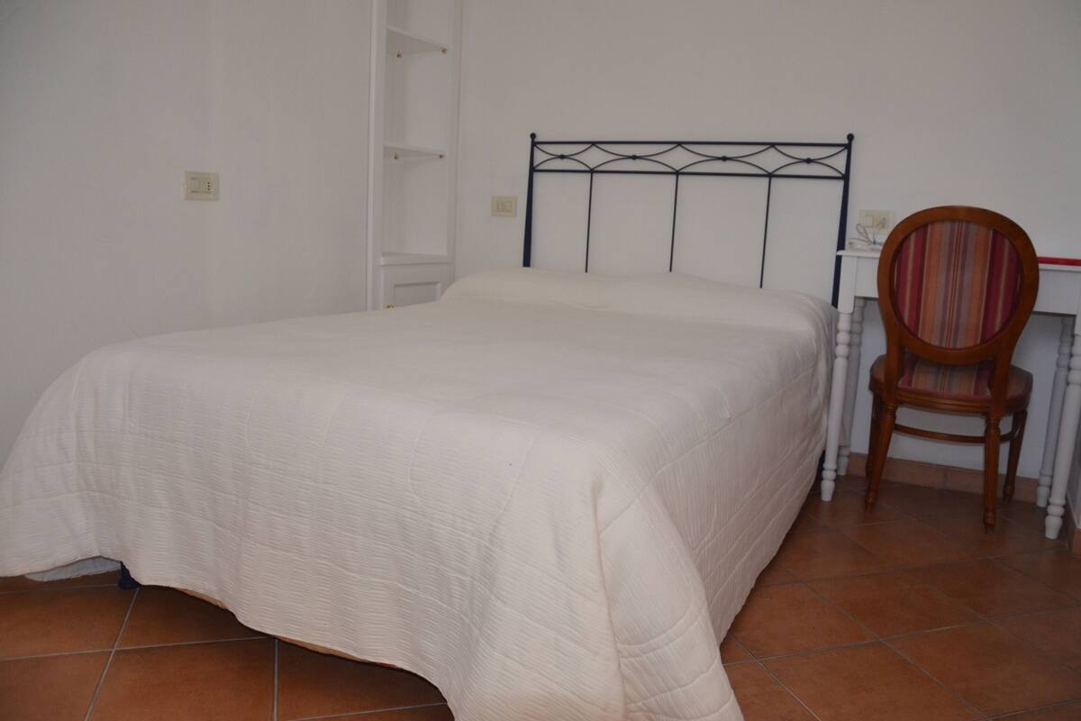 Una cama individual con cabecera de metal en una habitación con paredes blancas y suelo de baldosas.