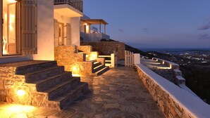 Una escalera de piedra conduce a un edificio blanco con contraventanas de madera, un balcón y vistas de un paisaje costero.