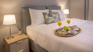 Un lit bien fait, avec du linge de lit blanc, un plateau de petit-déjeuner comprenant du jus, une tasse et une assiette avec une poire. Il y a deux lampes de chevet et une petite table de chevet avec une lampe et un vase.