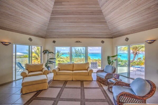A spacious living room with a high ceiling, large windows offering ocean views, and comfortable seating.
