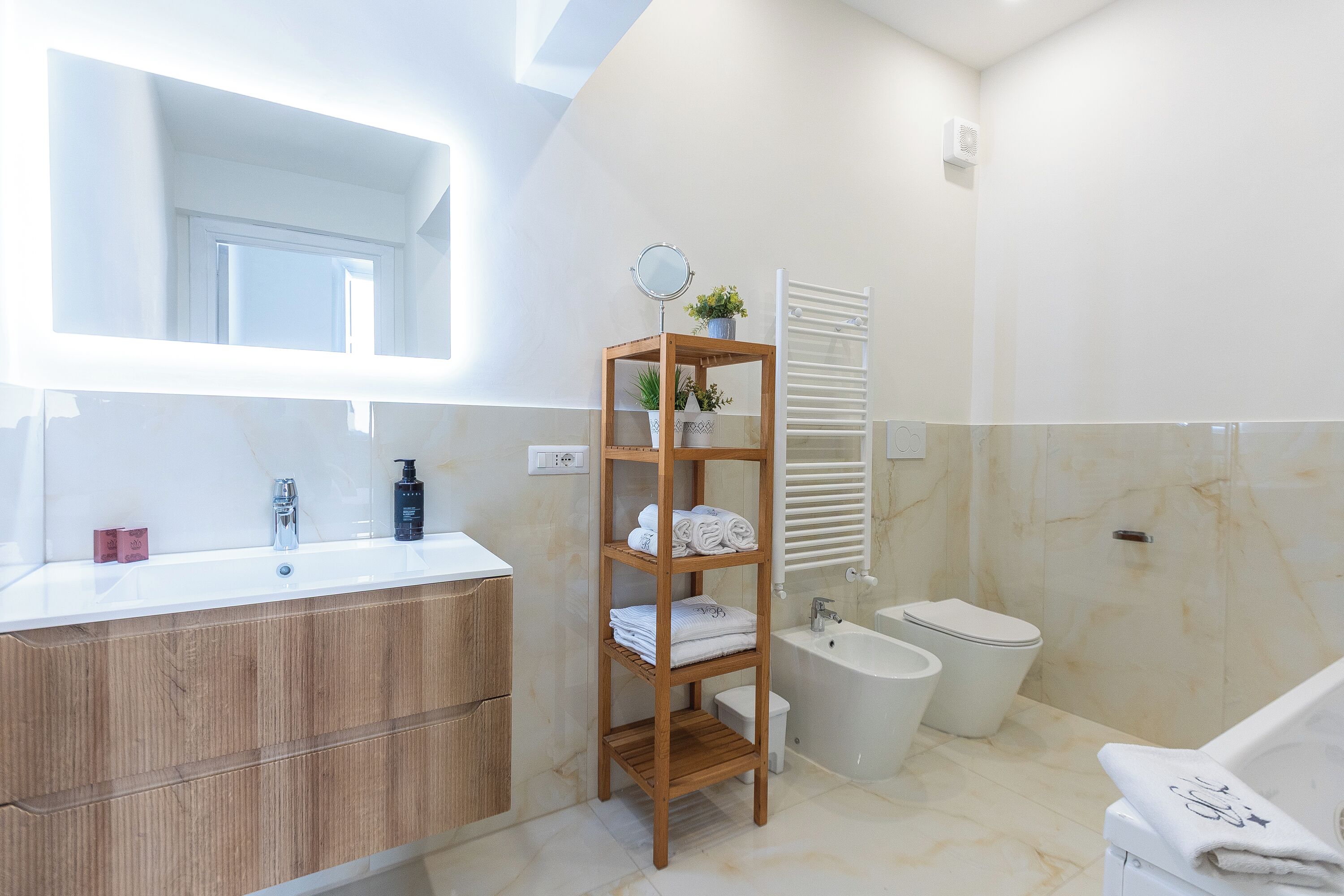 A modern bathroom with a wooden vanity, a mirror, a shelf with towels, a bidet, and a bathtub.