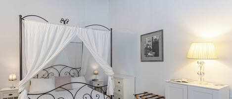 A four-poster bed with a canopy, a bedside table with a lamp, a small cabinet, and a wooden stool.