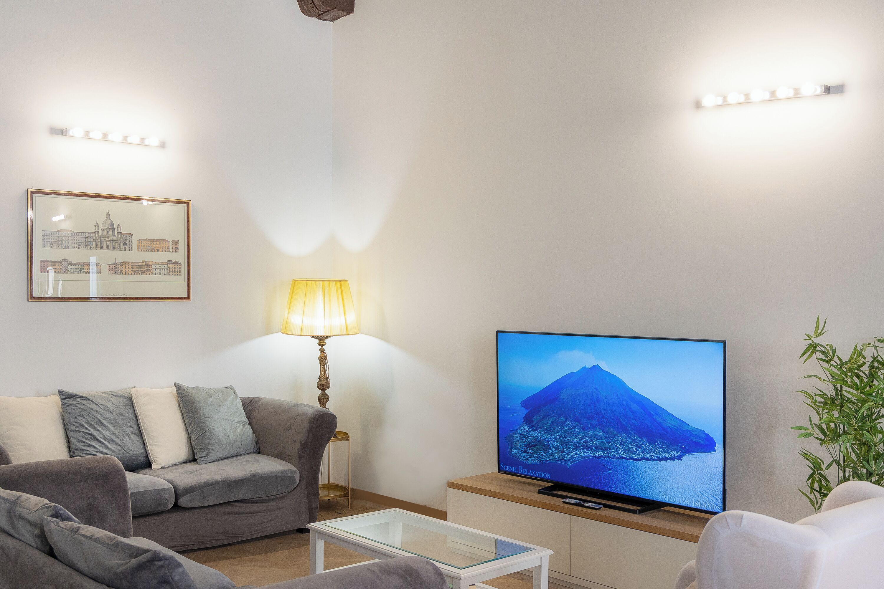 Ein Wohnzimmer mit grauem Sofa, einem Glastisch, einem Fernseher, der eine schöne Landschaft zeigt, einer Lampe und einem gerahmten Bild an der Wand.
