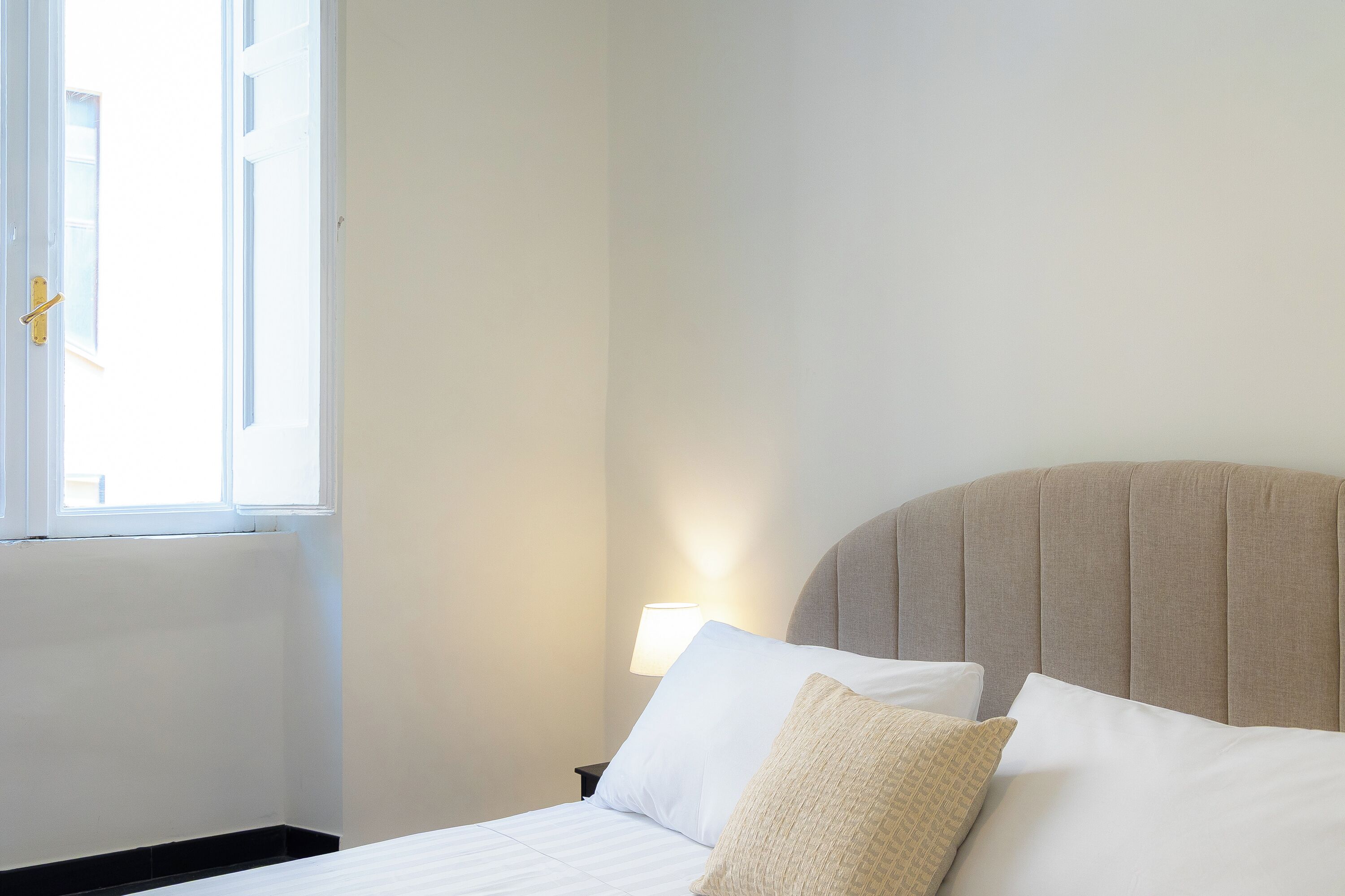 A neatly made bed with white linens and a beige headboard, a bedside lamp, and a window with a view of the outside.