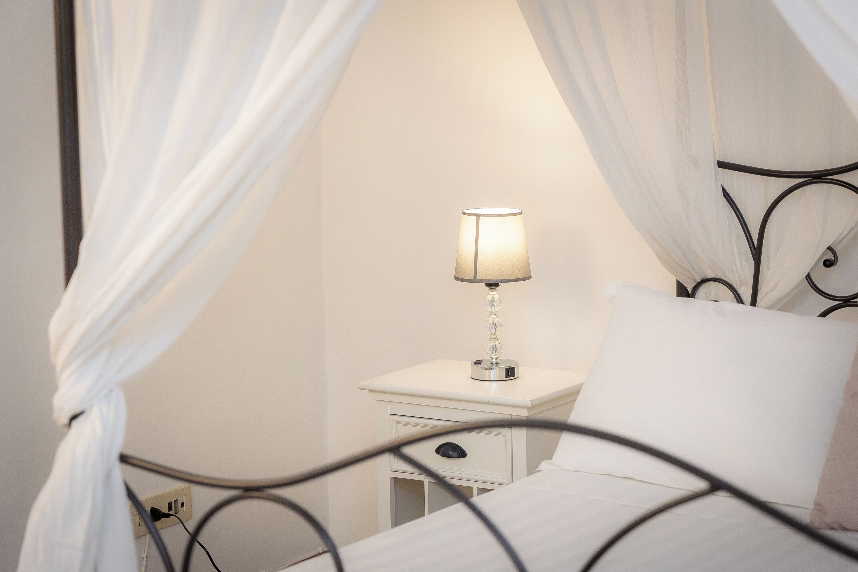 A bed with white linens, a bedside table with a lamp, and a power outlet.