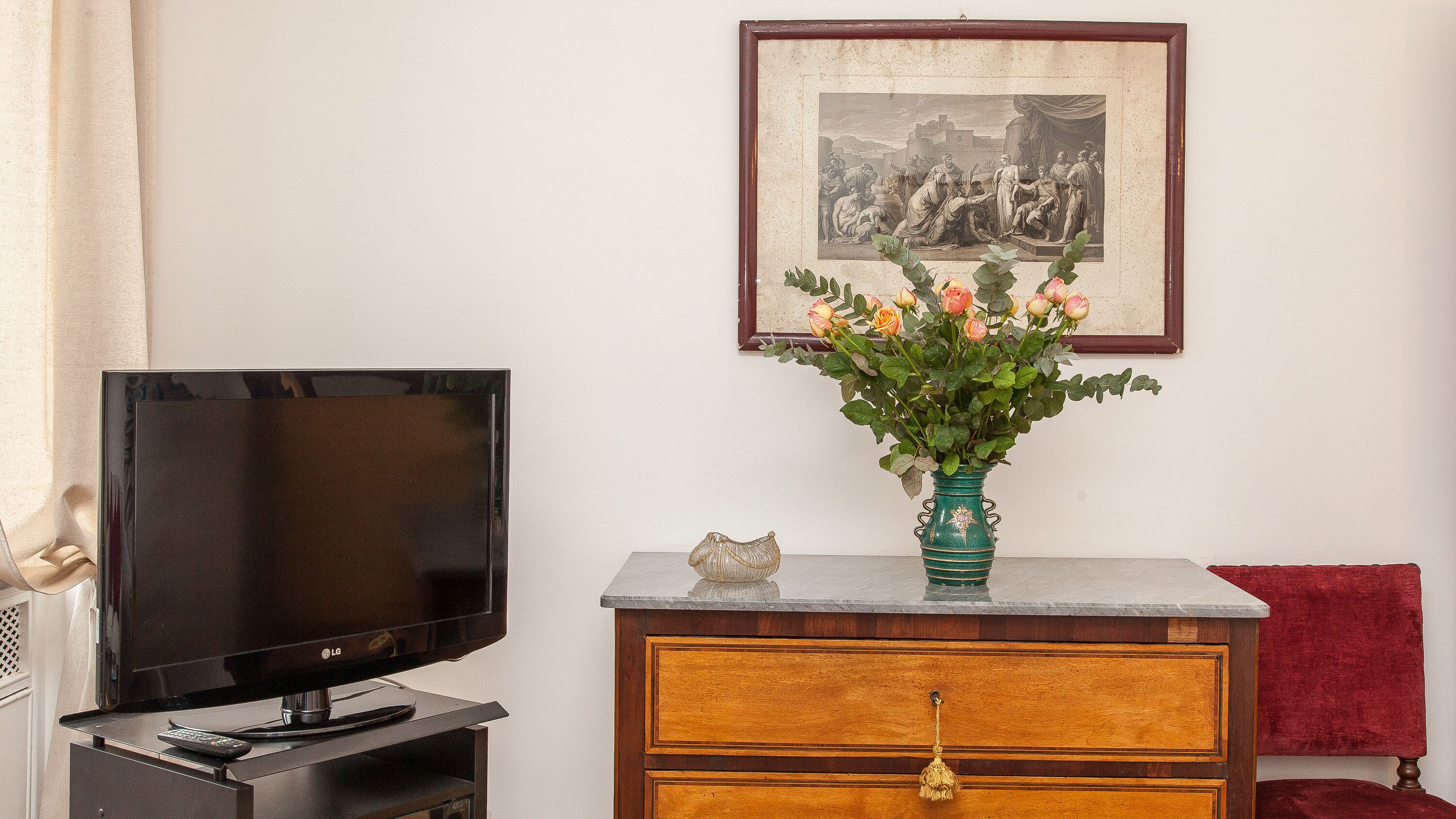 A living room with a flat-screen TV on a stand, a wooden cabinet with a vase of flowers, and a framed picture on the wall.