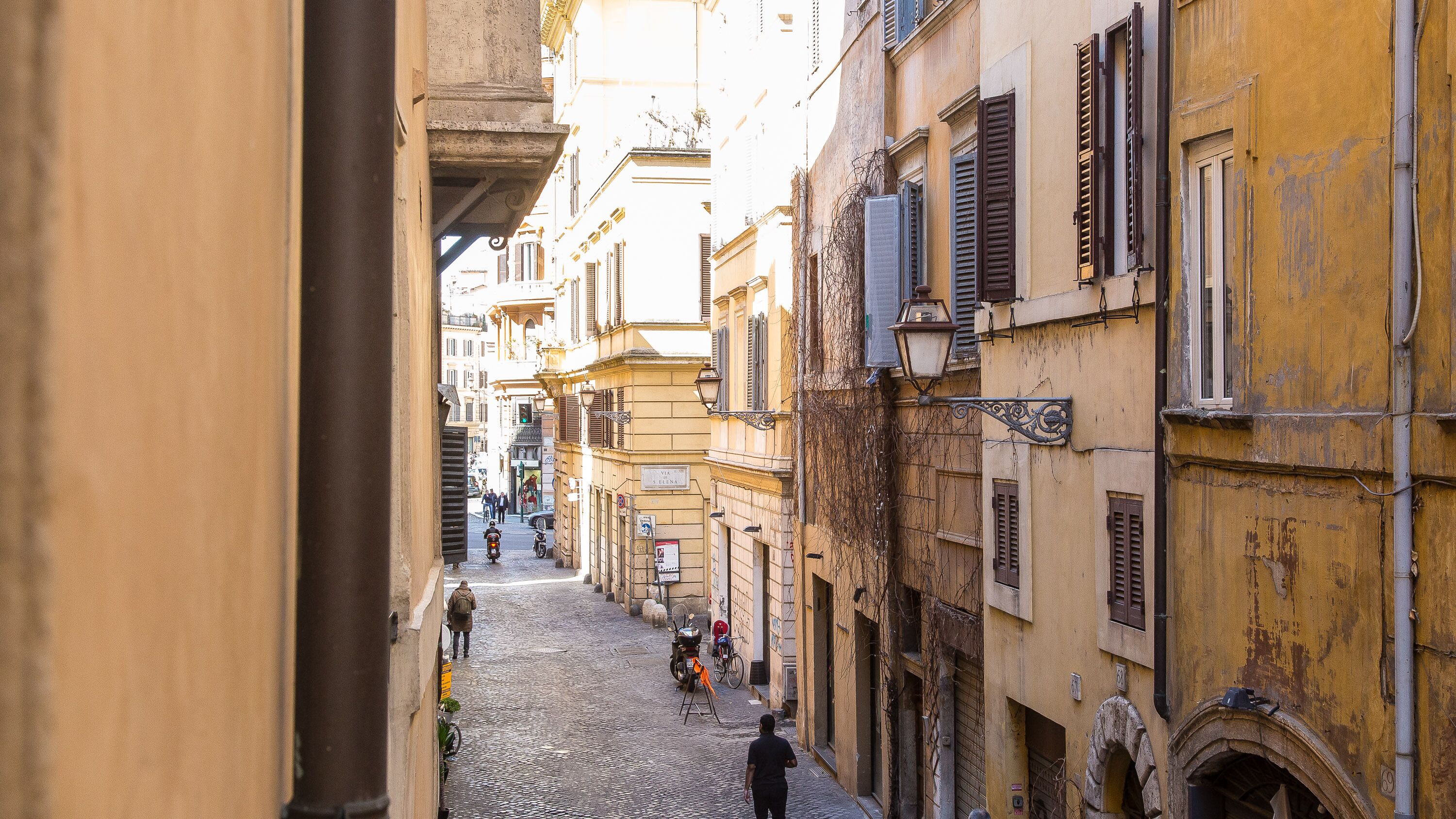 A narrow cobblestone street flanked by historic buildings with shuttered windows and a mix of architectural styles.