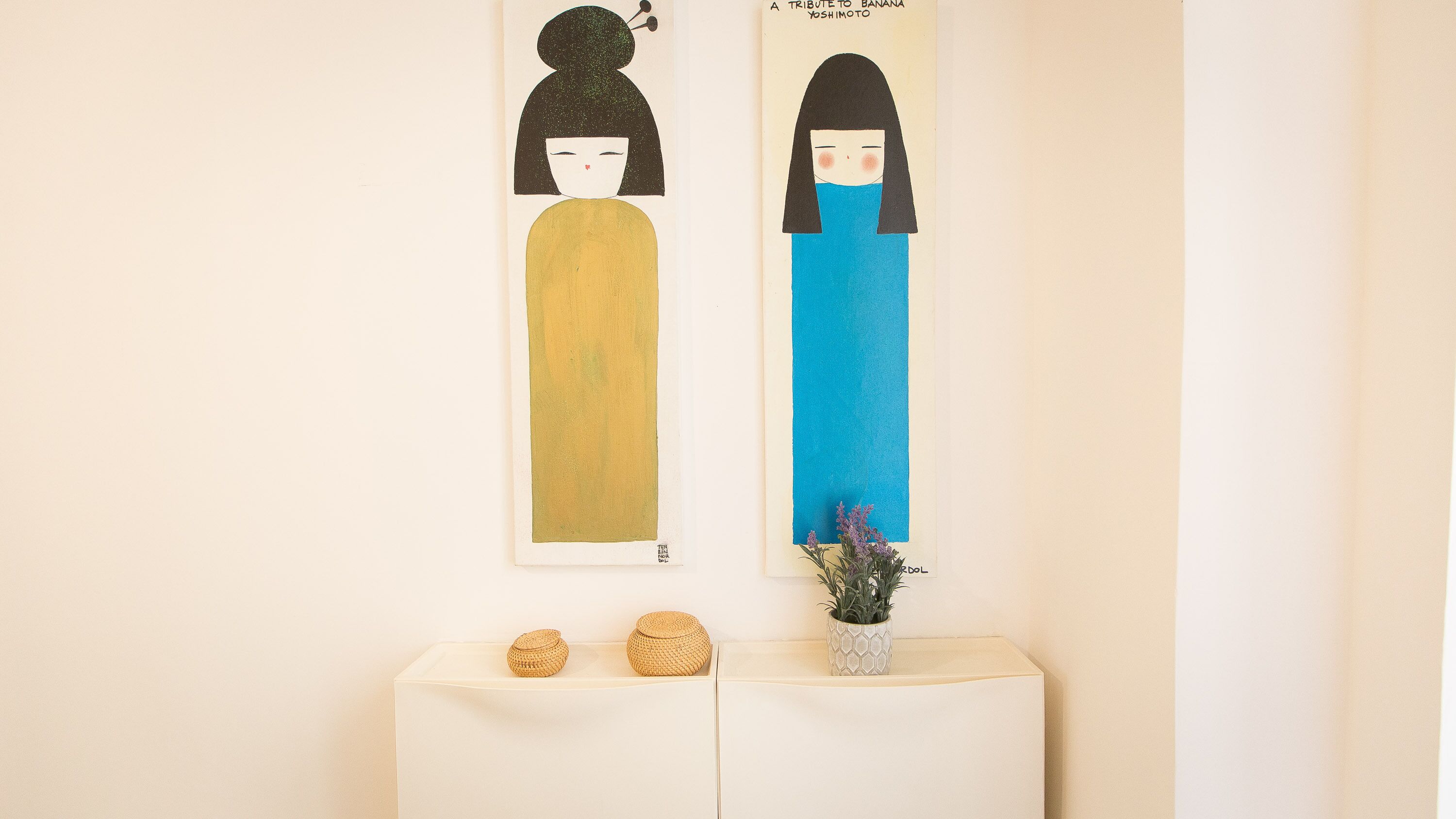 Deux œuvres d’art encadrées sont accrochées au mur, une petite table porte deux paniers en osier, et un pot de fleurs aux fleurs violettes est posé à proximité.