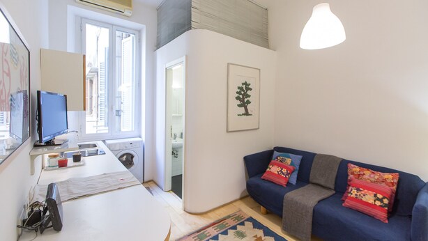 A compact living space with a washing machine, a flat-screen TV, a sofa with colorful cushions, and a framed artwork on the wall.