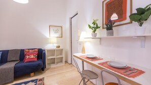 A small, cozy living room with a blue sofa, a dining table with chairs, a lamp, and a framed picture on the wall.