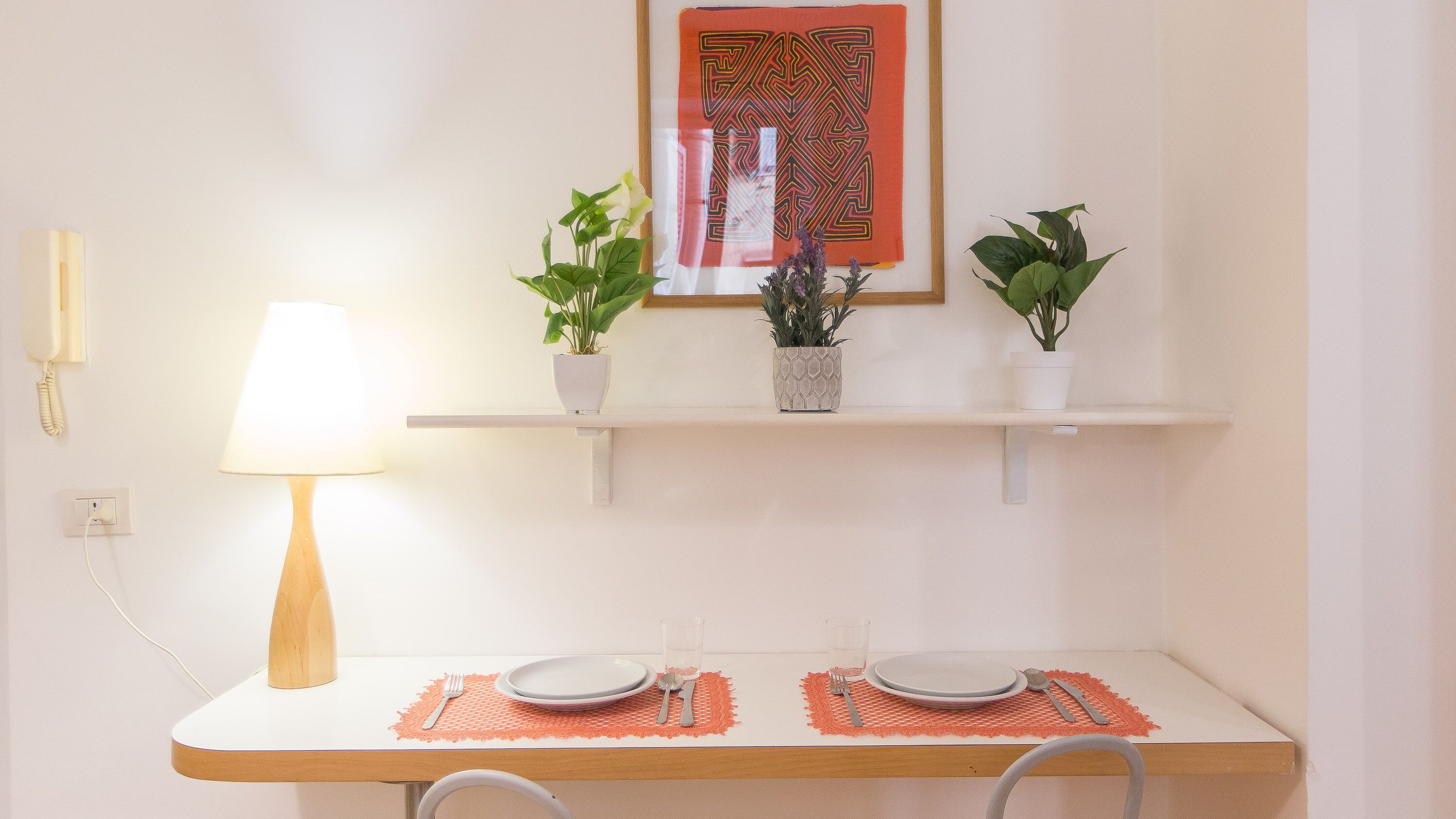 A dining area with a table set for two, a lamp, and two potted plants on a shelf above.