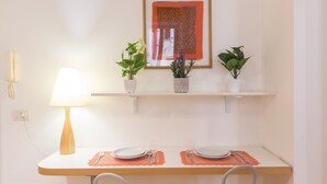 A dining area with a table set for two, a lamp, and two potted plants on a shelf above.
