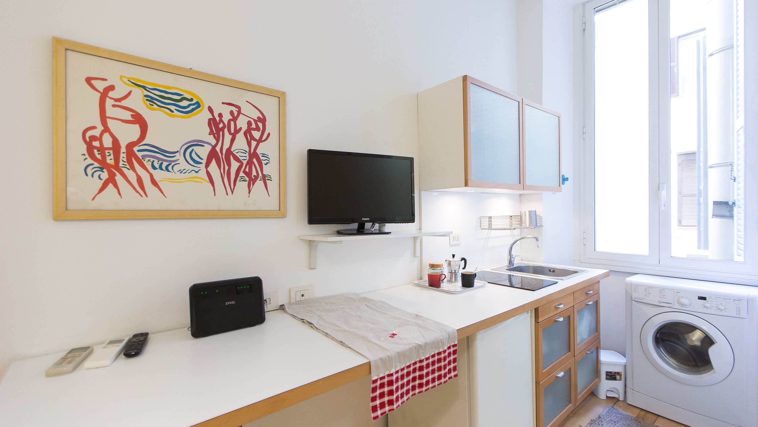 A small, modern kitchen with a flat-screen TV, a washing machine, and a window.