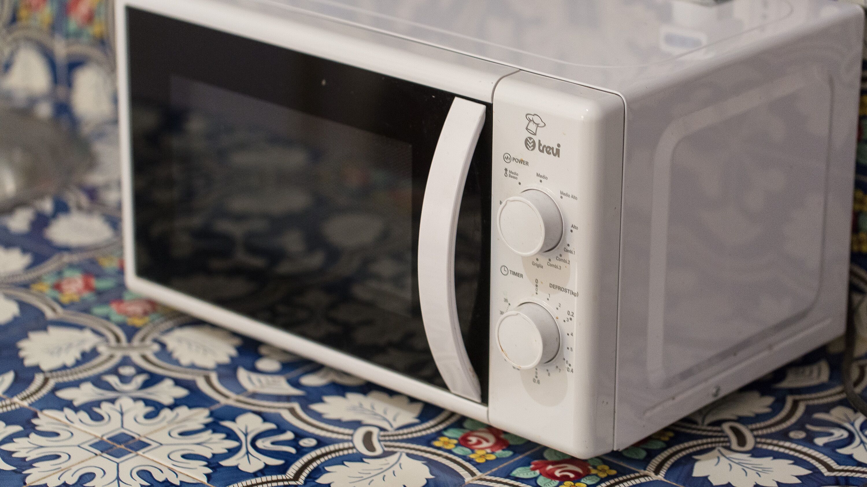 A white microwave oven with two control knobs and a digital display, placed on a patterned tablecloth.