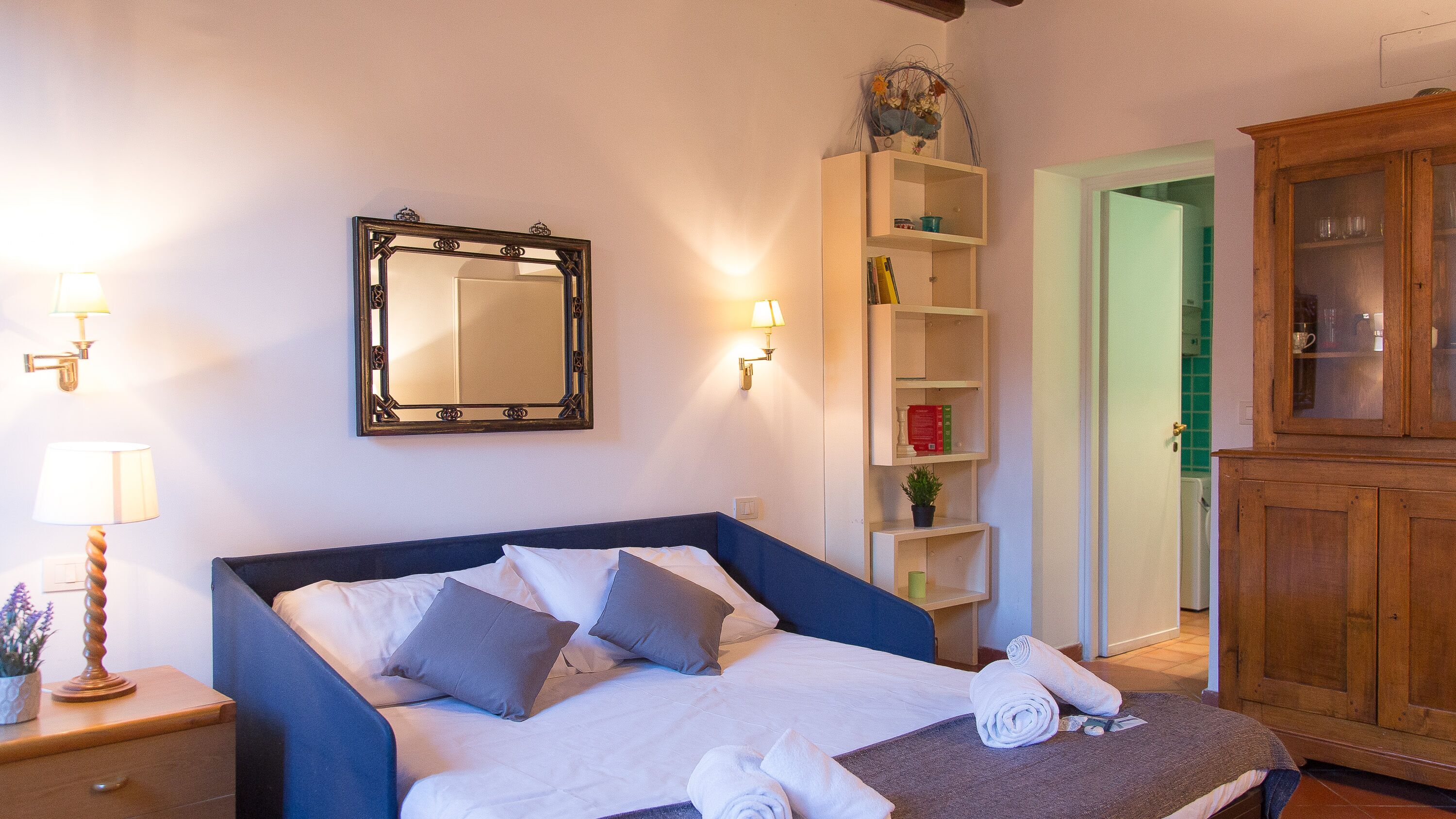 Una cama bien hecha con almohadas, una mesita de noche con lámpara, un espejo en la pared, un estante con libros y una planta, y un mueble de madera.