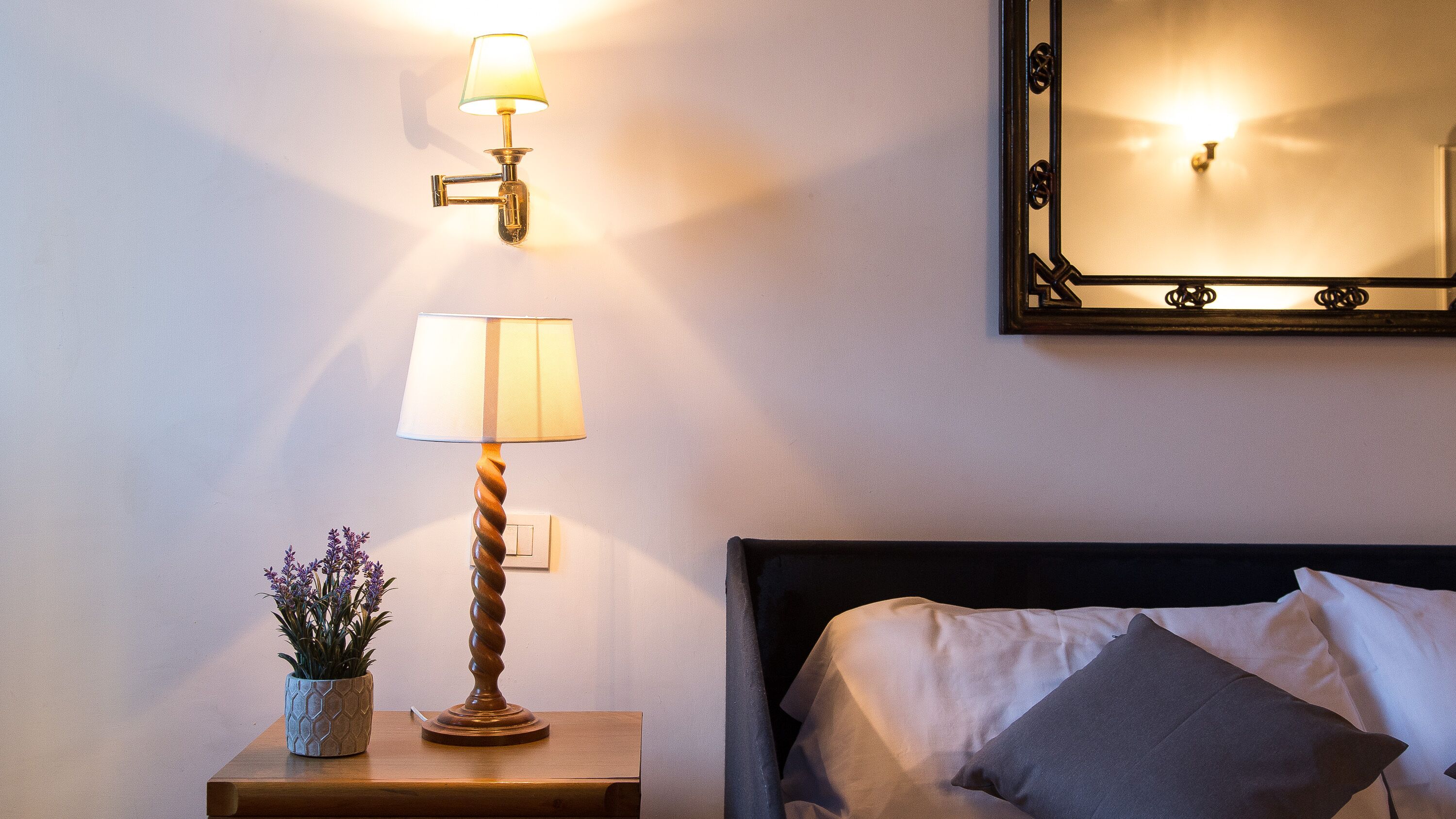 En pænt redt seng med puder, et natbord med lampe og en potteplante, samt et spejl på væggen.