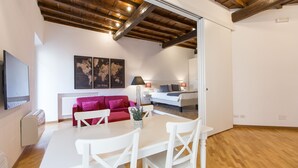 A modern living room with a wooden ceiling, a red sofa, a white dining table with chairs, and a bed with white bedding. - Rental in Rome Trastevere Atmosphere (Rome)