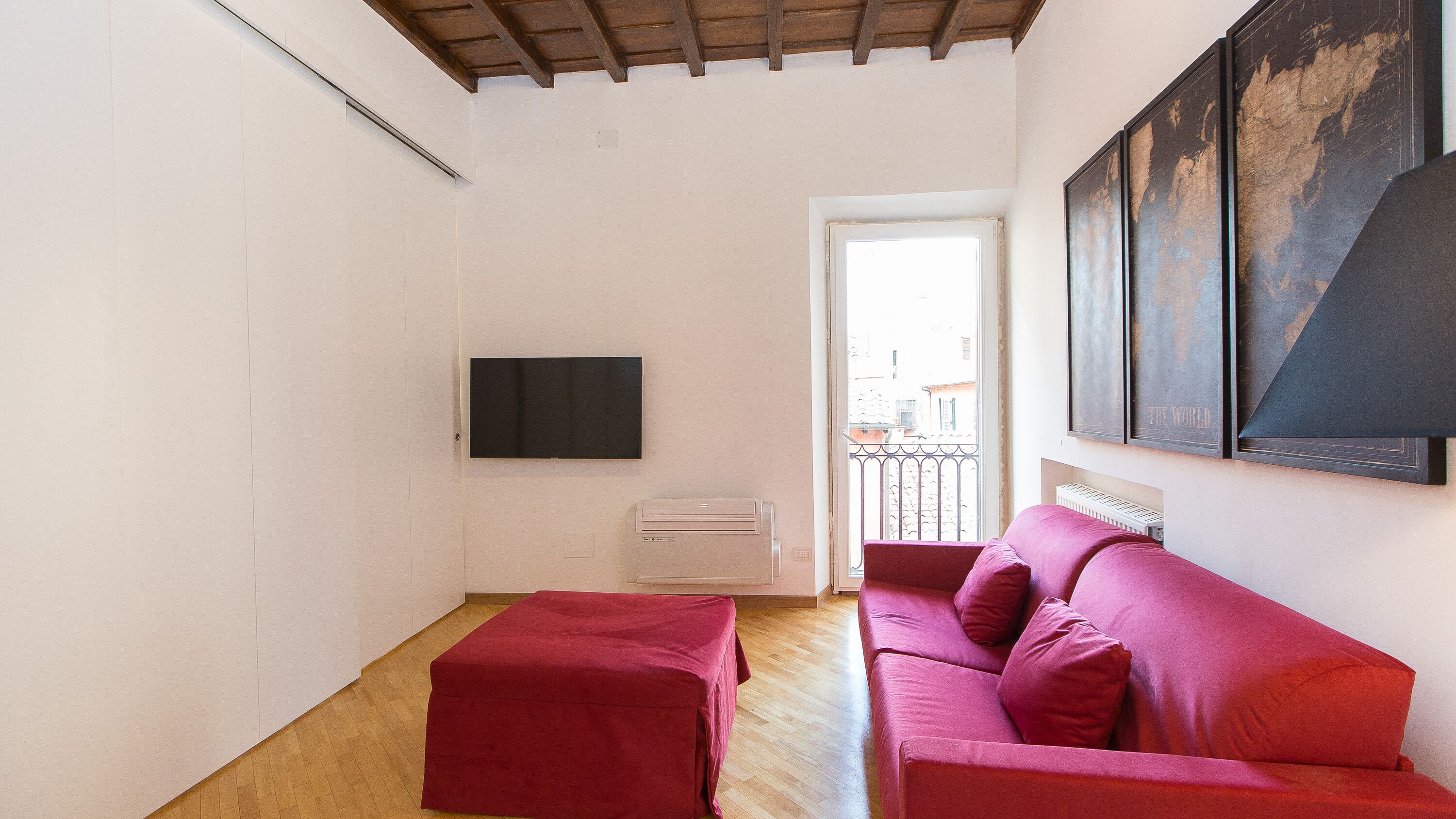 Una habitación con suelo de madera, un sofá rojo, un reposapiés rojo, un televisor de pantalla plana montado en la pared y una ventana con vistas a edificios.