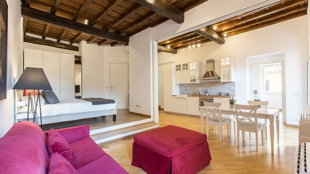 A modern interior with a bed, a red ottoman, a dining area with a table and chairs, and a kitchen with white cabinets and a stove. - Rental in Rome Trastevere Atmosphere (Rome)