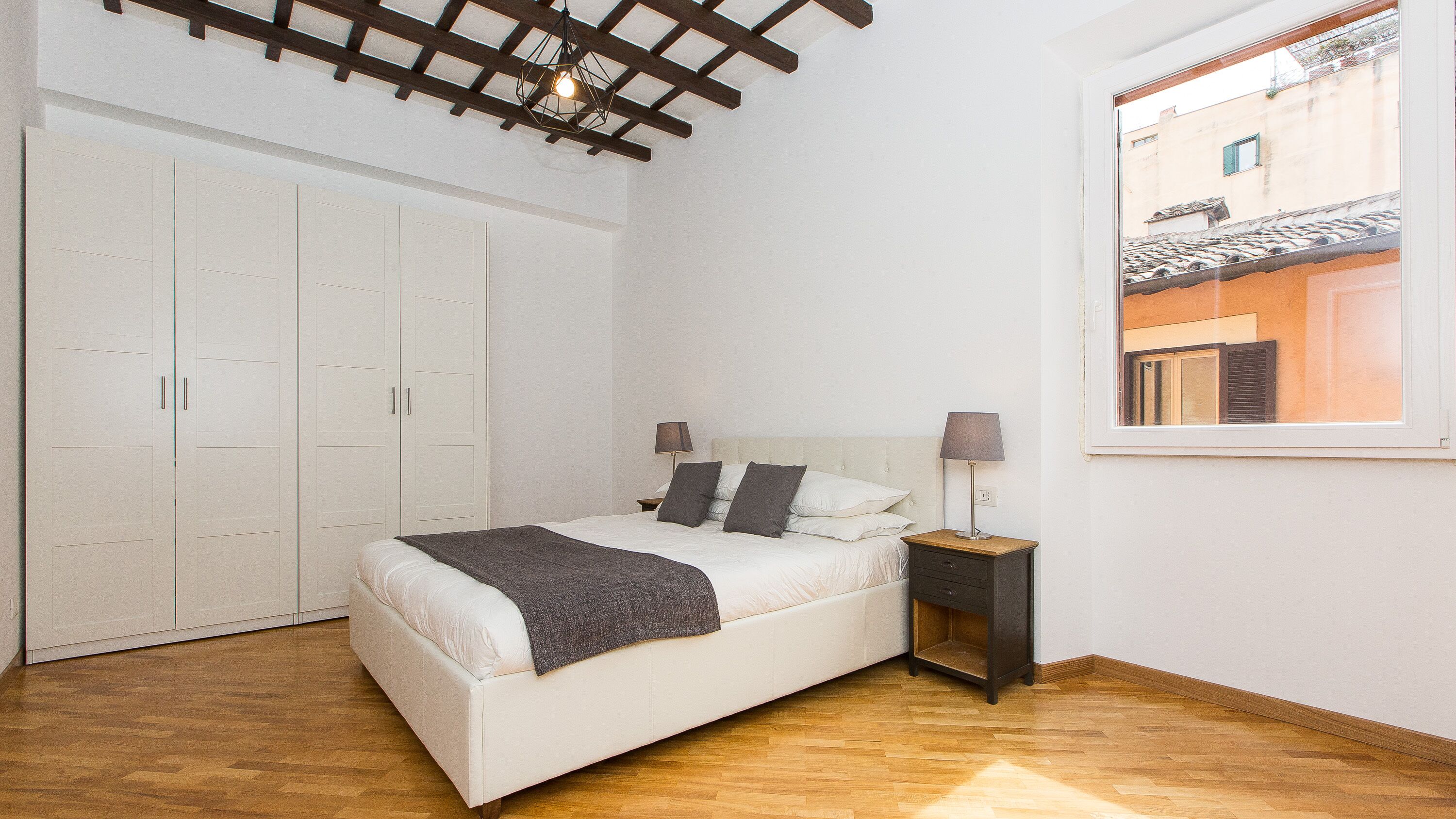 Un dormitorio con cama, mesitas de noche, armario y ventana con vistas a edificios.