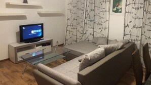 A modern living room with a glass coffee table, a grey sofa, a flat-screen TV on a white stand, and a wall with a tree branch pattern curtain. - Apartmán Karla Čapka (Teplice)