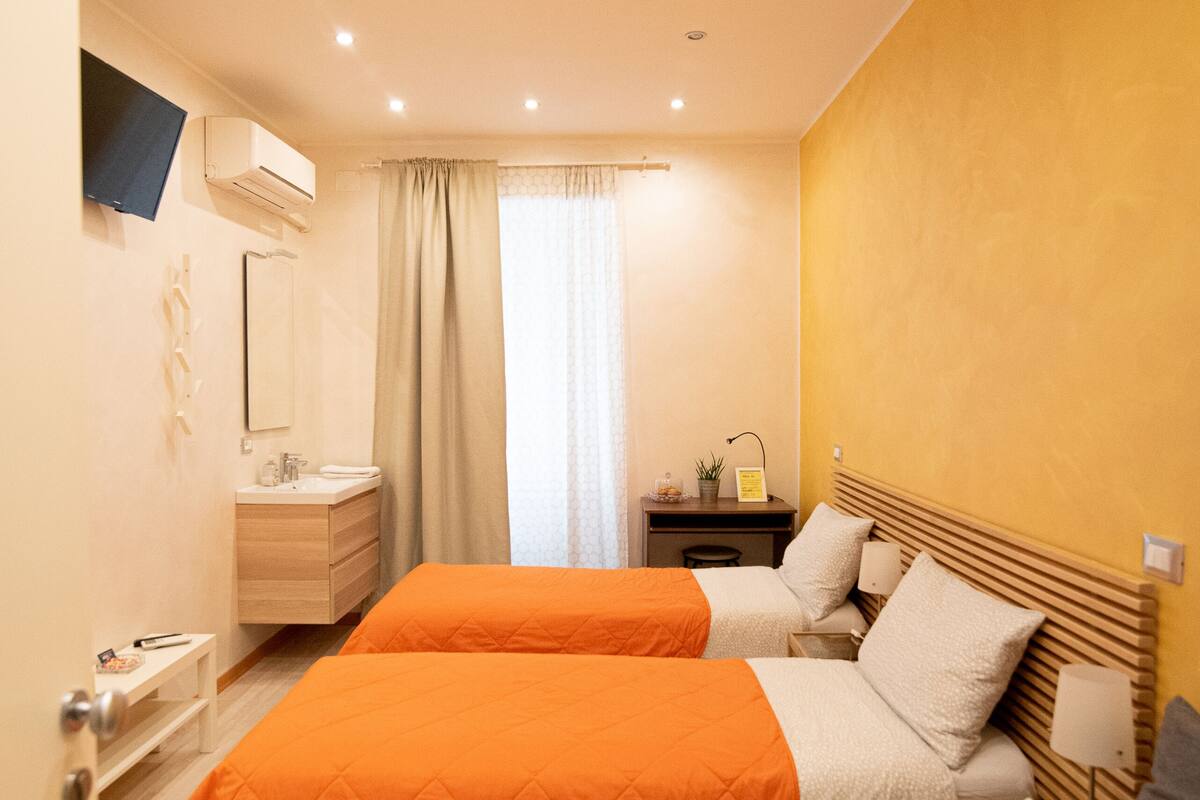 Une chambre d’hôtel avec deux lits, une table de chevet, une commode, une télévision, un miroir, une fenêtre avec des rideaux et un plancher en bois.