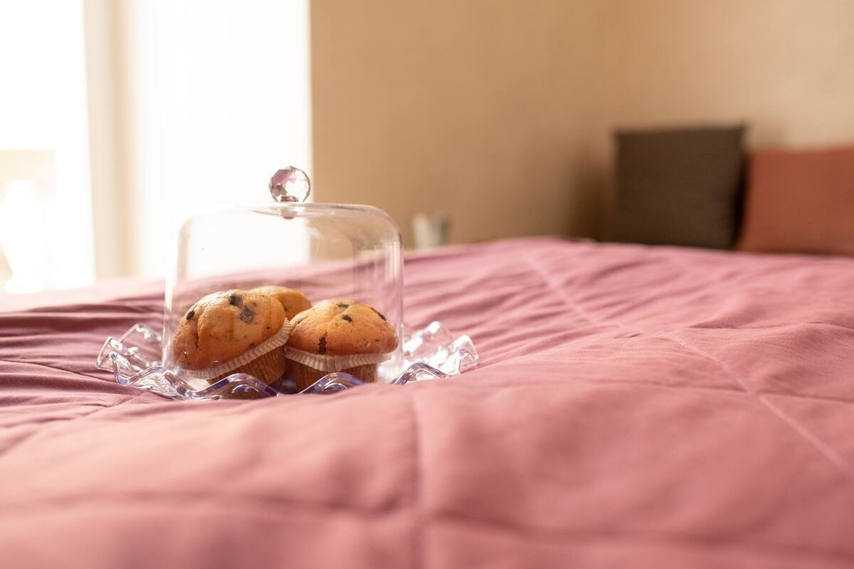 Un lit recouvert d’une couverture rose et surmonté d’une cloche en verre transparent contenant des muffins.