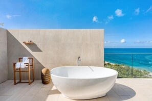 A modern bathroom with a freestanding bathtub, a wooden chair with towels, and a view of the ocean.