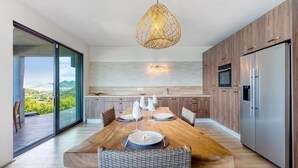 A modern kitchen with a wooden dining table set for four, a large window with a view of greenery, and a hanging light fixture. - Villa Amandara (Les Terres Basses)