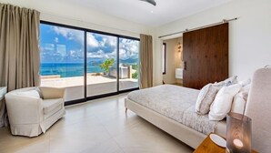 A bedroom with a large bed, a chair, a ceiling fan, and a view of the ocean through a sliding door. - Villa Amandara (Les Terres Basses)