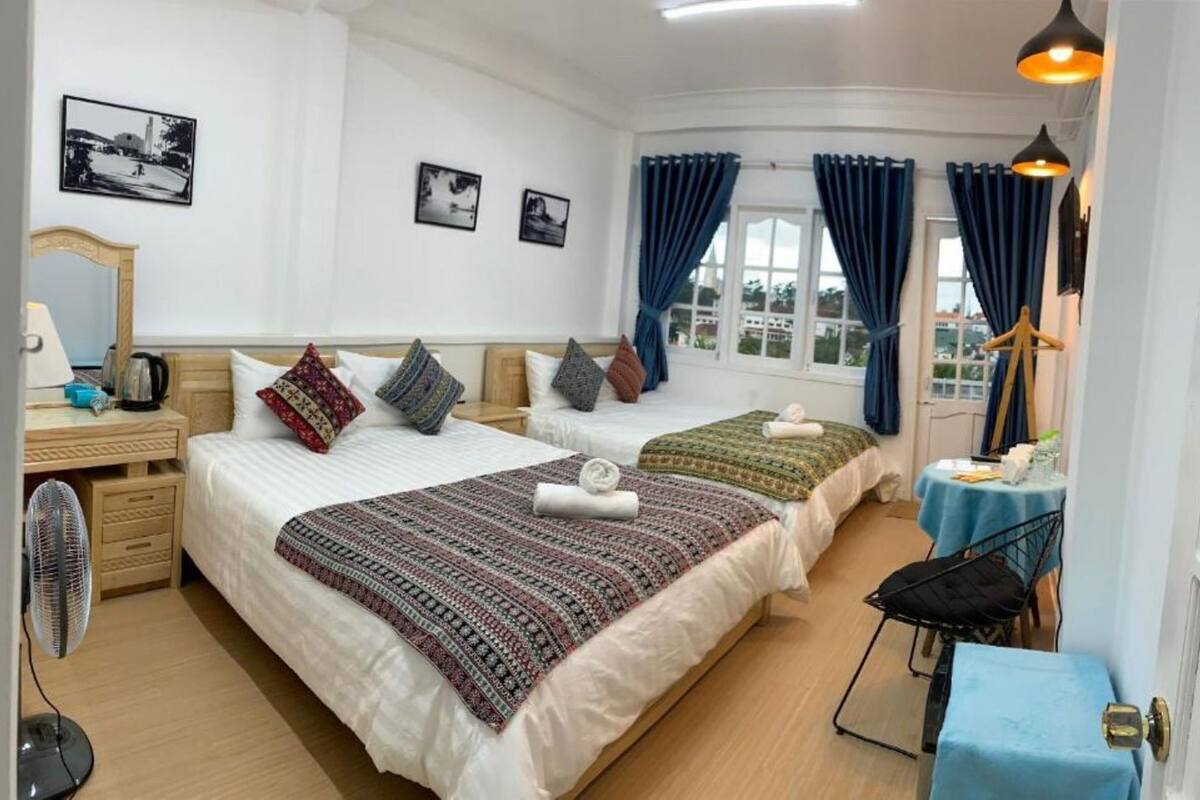 A hotel room with two beds, a desk, a chair, a table, a fan, and a window with curtains.