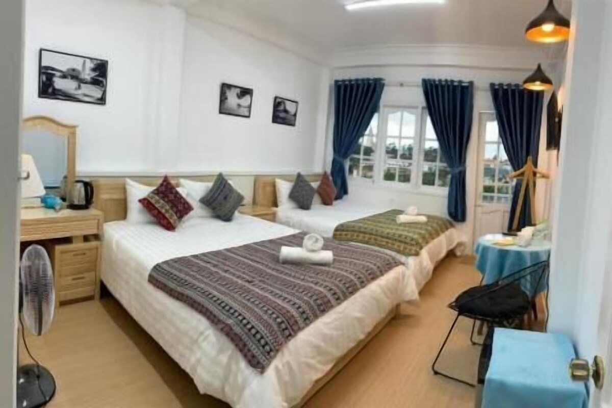Habitación de hotel con dos camas, un escritorio, una silla, una mesa, un ventilador y una ventana con cortinas.