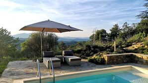 Una piscina con plataforma de piedra, dos sillas de descanso y una sombrilla.