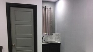 A small, compact bathroom with a grey door, a sink with a mirror above it, and a cabinet with a white countertop. - Island Vibes Suites (Port of Spain)