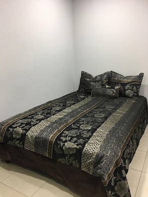 A bed with a dark floral-patterned bedspread and matching pillows in a room with plain white walls and tiled flooring. - Island Vibes Suites (Port of Spain)