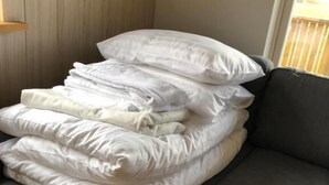 Una esquina de una habitación con un sofá gris, una pila de almohadas blancas y un piso de madera.