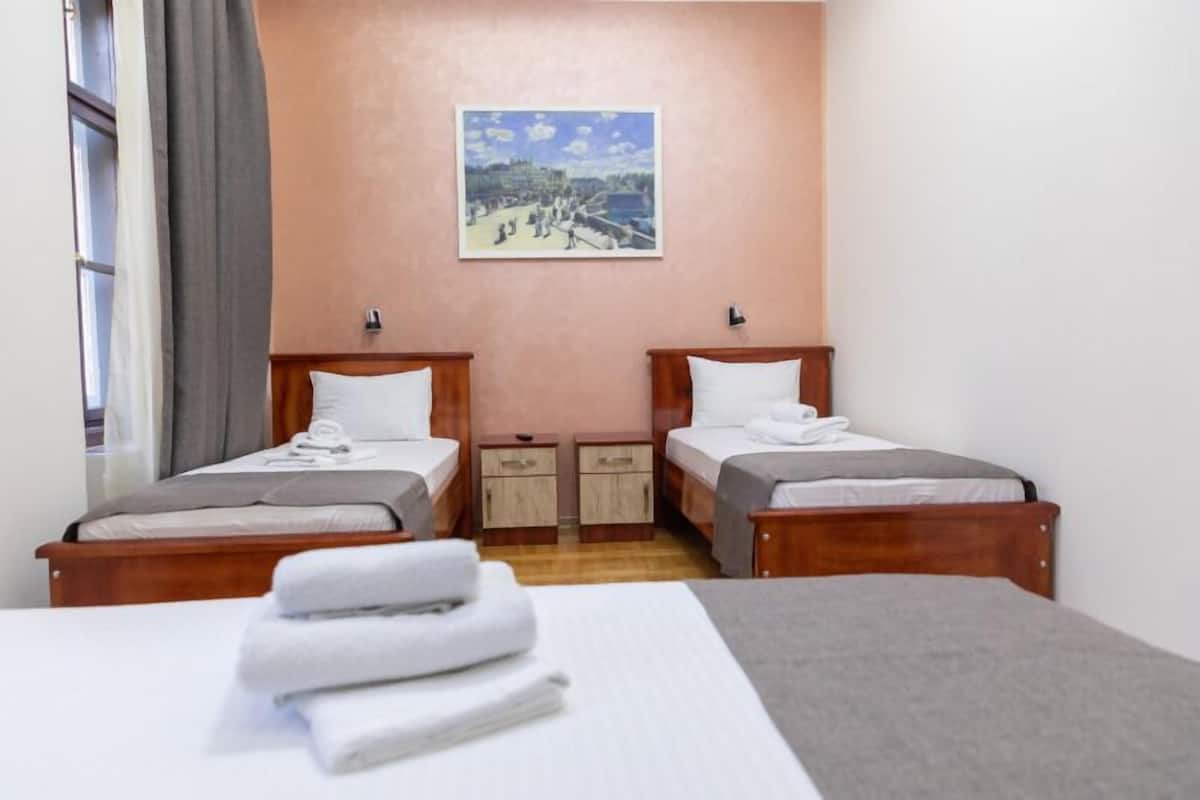 A hotel room with two single beds, a large bed, a painting on the wall, and a window with curtains.