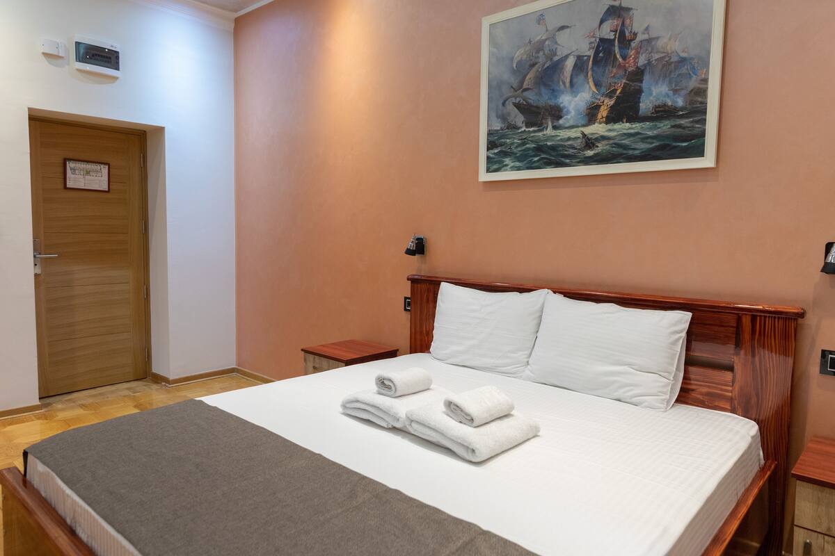 A neatly made bed with white linens and rolled towels, a wooden headboard, a painting of a ship on the wall, a wooden door, and a small bedside table.