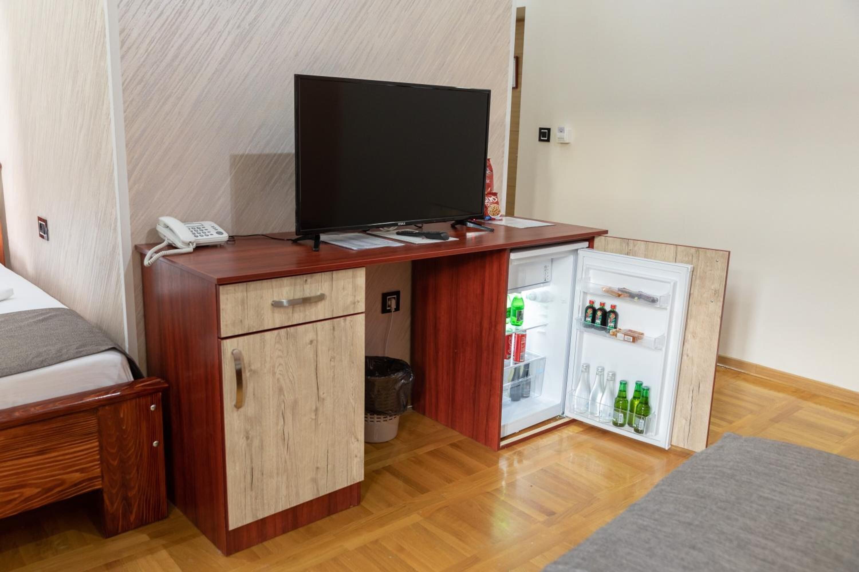 Une chambre compacte comprenant un bureau en bois, une télévision, un mini-réfrigérateur rempli de boissons, une petite poubelle et un lit avec une couverture grise.