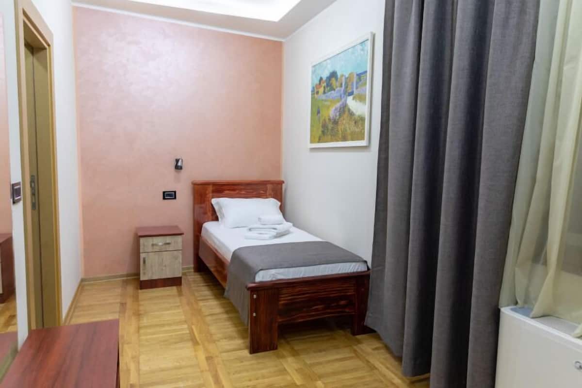 A bedroom with a wooden bed, a bedside table, a small cabinet, a painting on the wall, and a window with curtains.