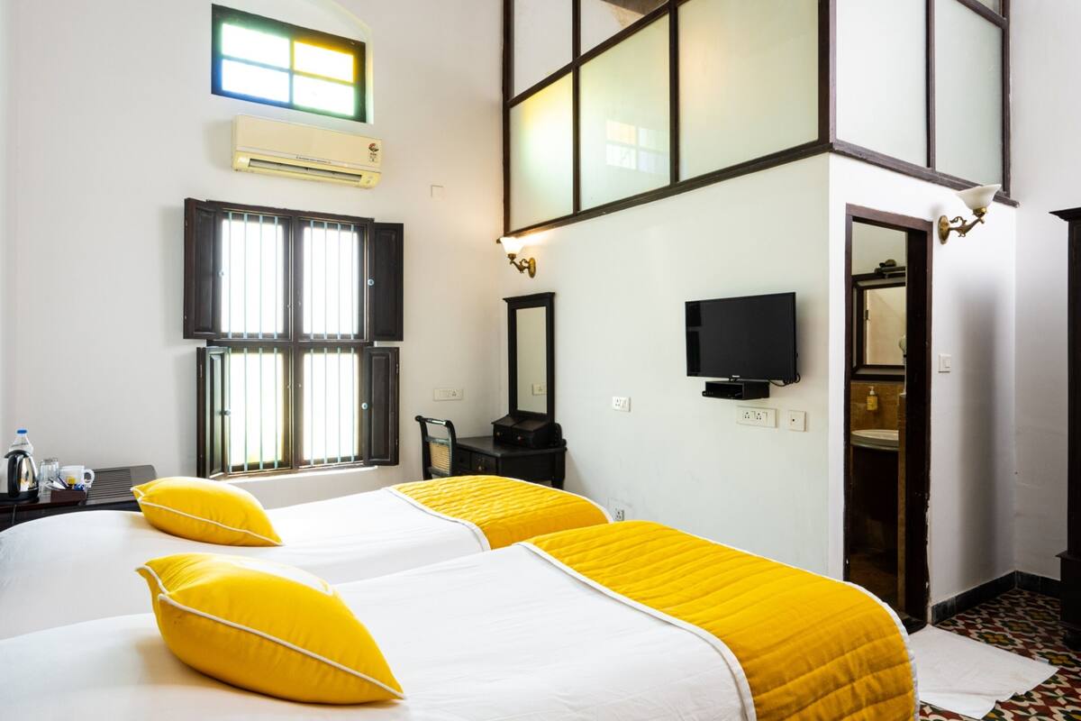 A hotel room with two beds, a television, an air conditioning unit, and a window with blinds.
